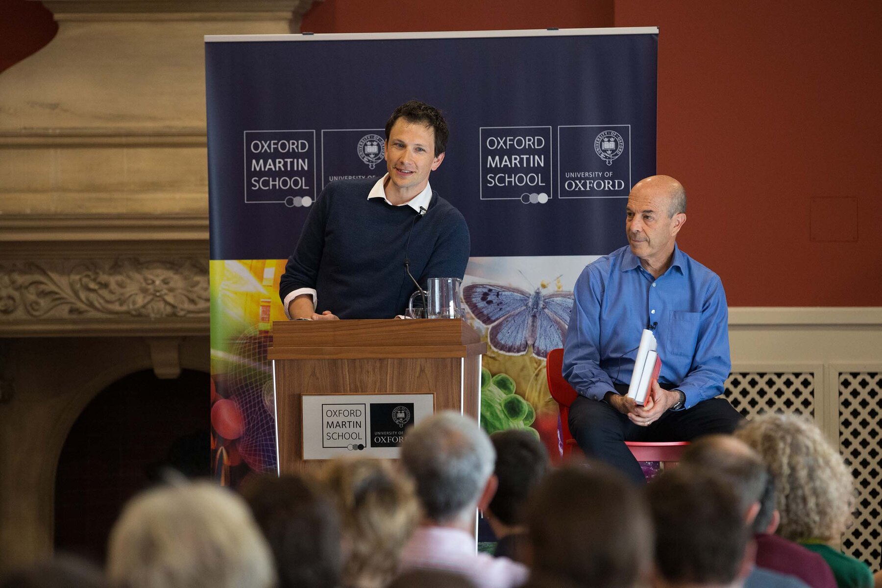 Chris speaking at a podium in front of Oxford Martin School banners, Ian Goldin seated beside him holding the book, the backs of a full audience in front