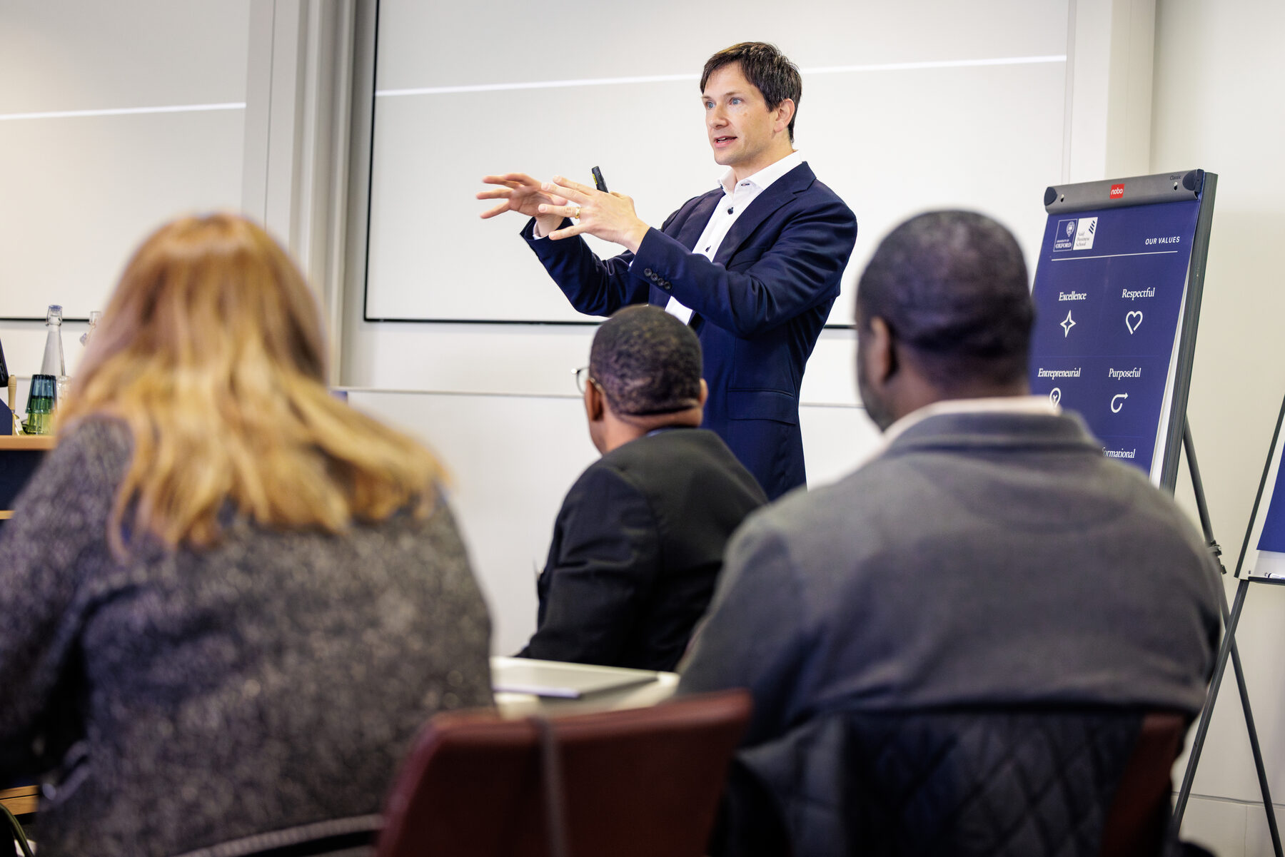 Chris Kutarna mid-gesture in an Oxford classroom, seen over the shoulders of three participants